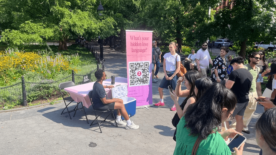 Micah at a folding table in Washington Square Park with a pink 'What's your hidden love language?' banner and a crowd